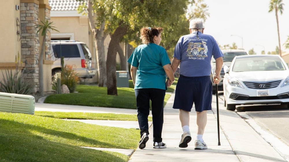 Senior male client using a cane is walking down the street holding hands with his female Right at Home caregiver