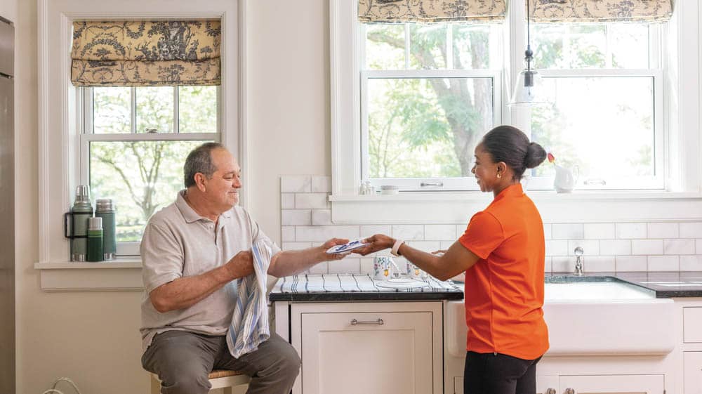 A female Right at Home caregiver is doing the dishes in a kitchen. She is handing the wet dish to a senior male client to dry the dish.