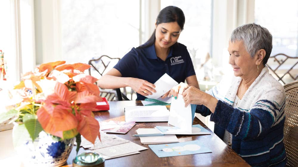 Female Right at Home caregiver helping senior female client stuff holiday cards into envelopes at a kitchen table 