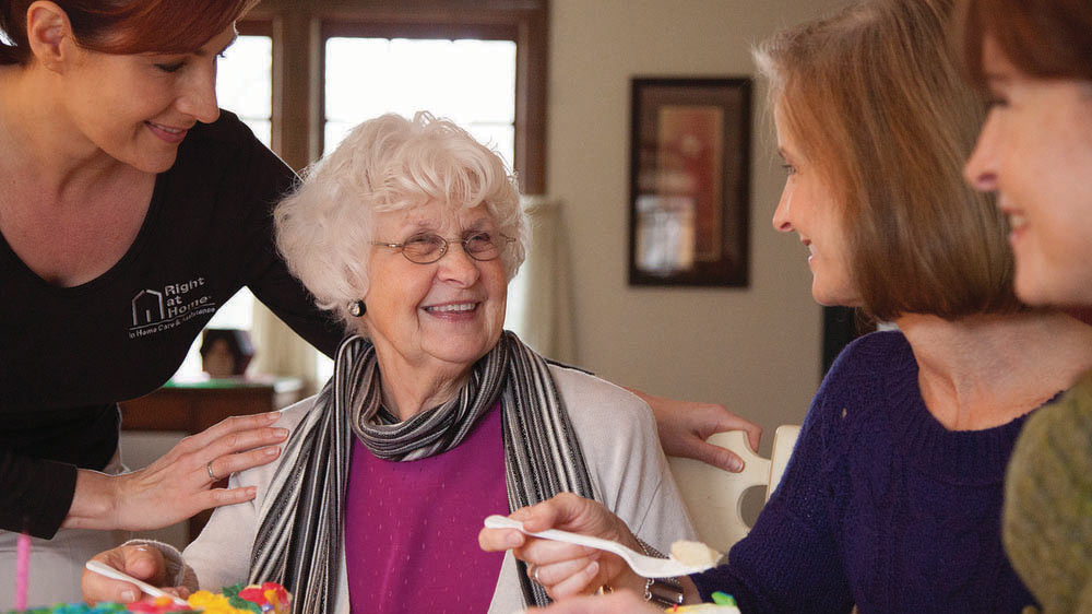 A female senior at the kitchen table with her two adult daughters and a female Right at Home caregiver, eating birthday cake