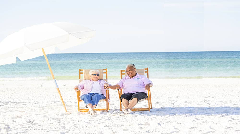 Female caregiver and female senior sitting on a beach with a beach umbrella 