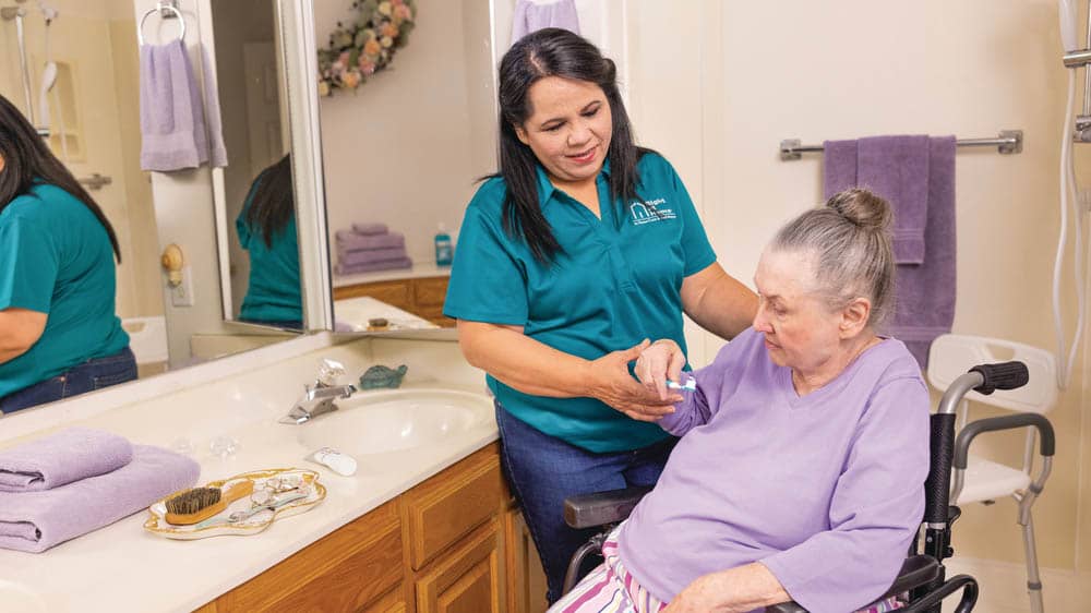 A female caregiver is helping a senior female in a wheelchair brush her teeth
