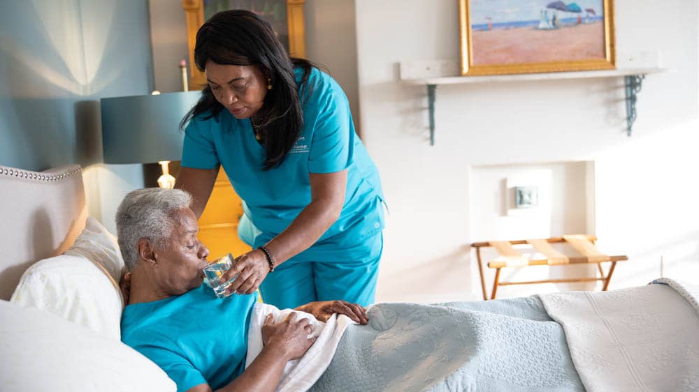 A female Right at Home caregiver is helping a senior male client drink a glass of water while he is laying in bed