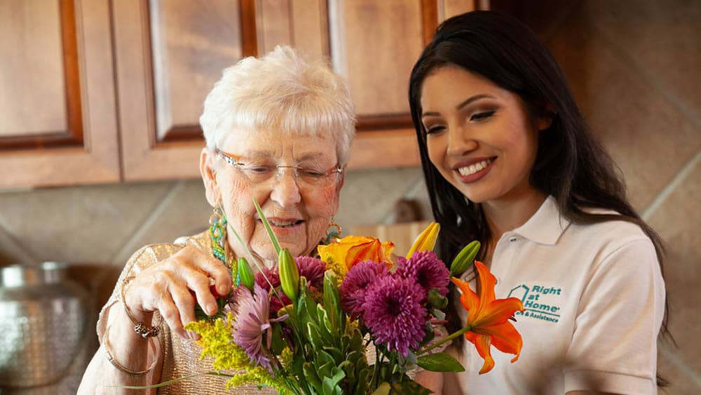 A female Right at Home caregiver helps a senior female client with a flower arrangement in the kitchen