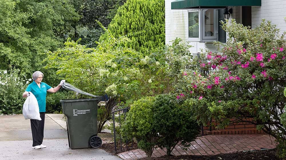 A female Right at Home caregiver is throwing a trash bag into a trash bin outside of a house