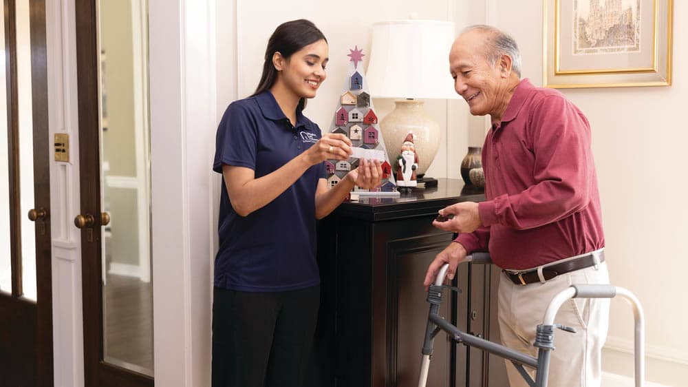 A female Right at Home caregiver is holding up a piece of paper for a senior male client to read, while standing with his walker inside at Christmas time.