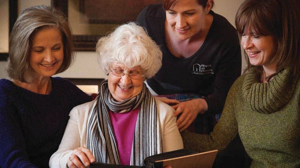 A female senior is looking at photo album with her two adult daughters and a female caregiver from Right at Home