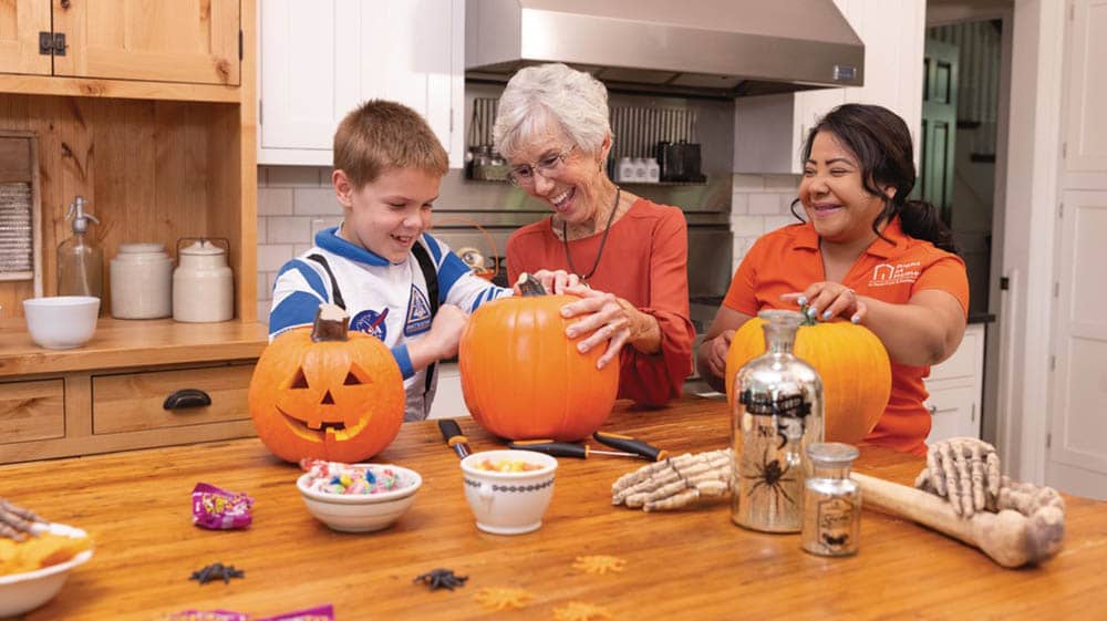 A senior female client is carving a pumpkin with her grandson at a kitchen table, while a female Right at Home caregiver looks on