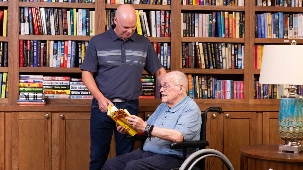 A male caregiver is standing next to a senior male client who is sitting in a wheelchair, looking at a book in a library