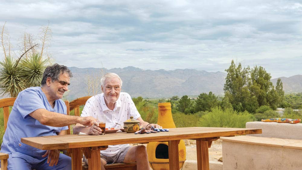 A male caregiver from Right at Home helping a senior male eat while sitting outside at a table