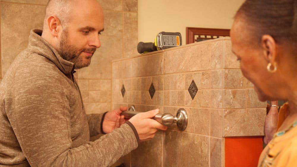 A man is installing a safety hand bar in a shower while a female senior client looks on