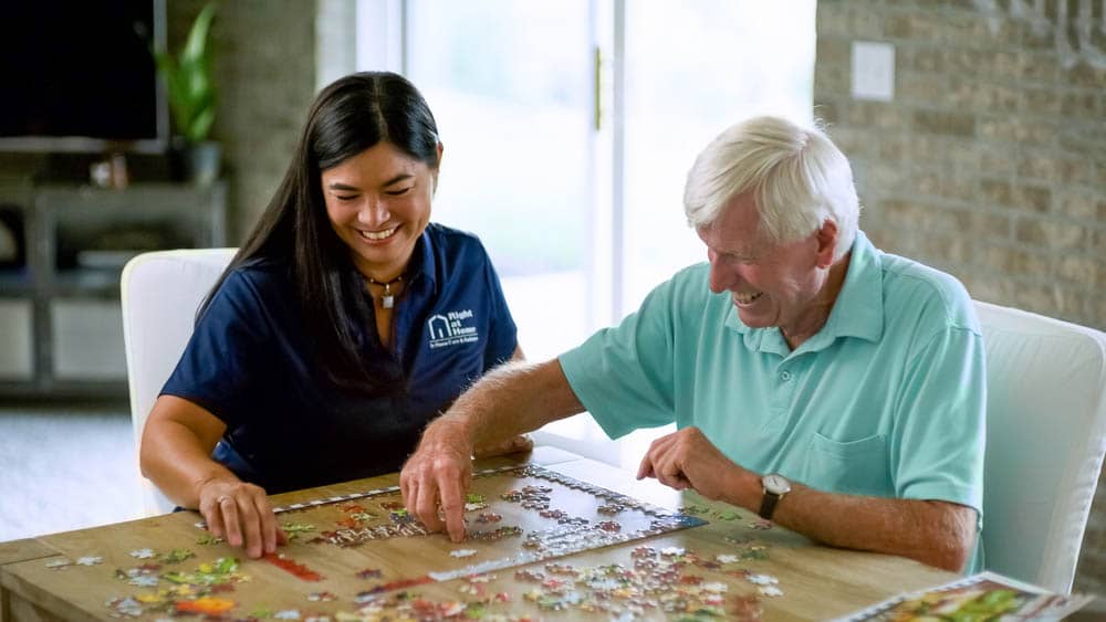 Female Right at Home caregiver sitting at a table with a senior male client putting a puzzle together