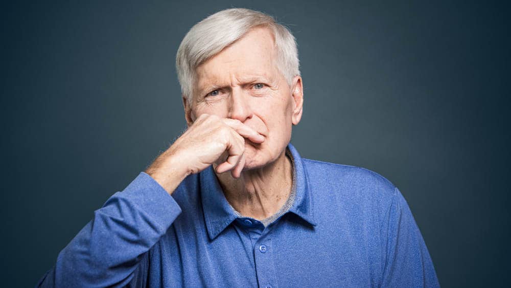 A photo of a senior male with a look of concern on his face and hand near mouth in front of a gray background