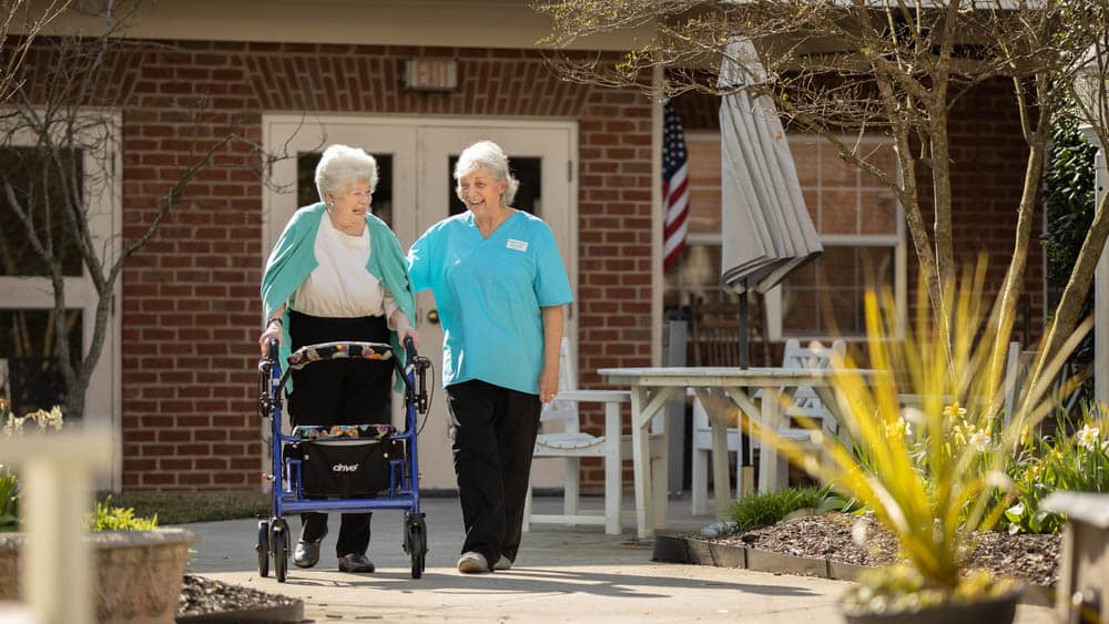 Female Right at Home caregiver is helping a senior female client walk outside. The senior female client is using a walker for additional assistance