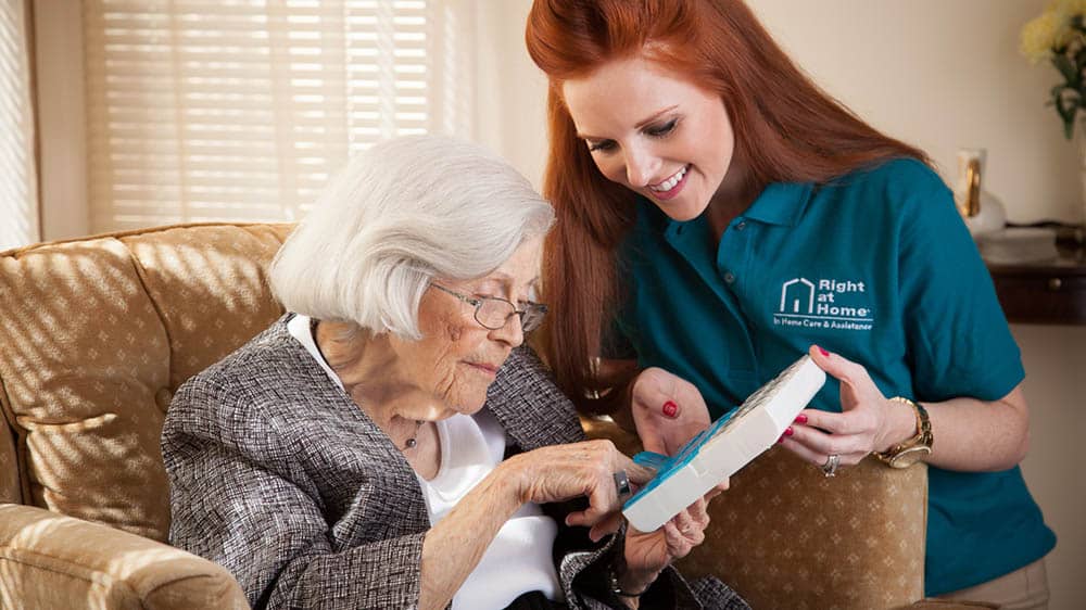 Female Right at Home caregiver helping a senior female client with a pill dispenser while client sits in armchair. 