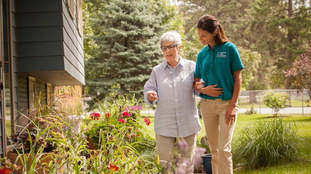 Female Right at Home caregiver and female senior client are walking arm in arm outside while looking at flowers