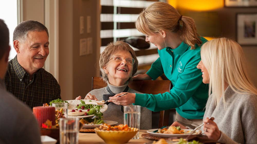 Family sitting at a table while a Right at Home caregiver is helping serve food to a senior female sitting with the family.
