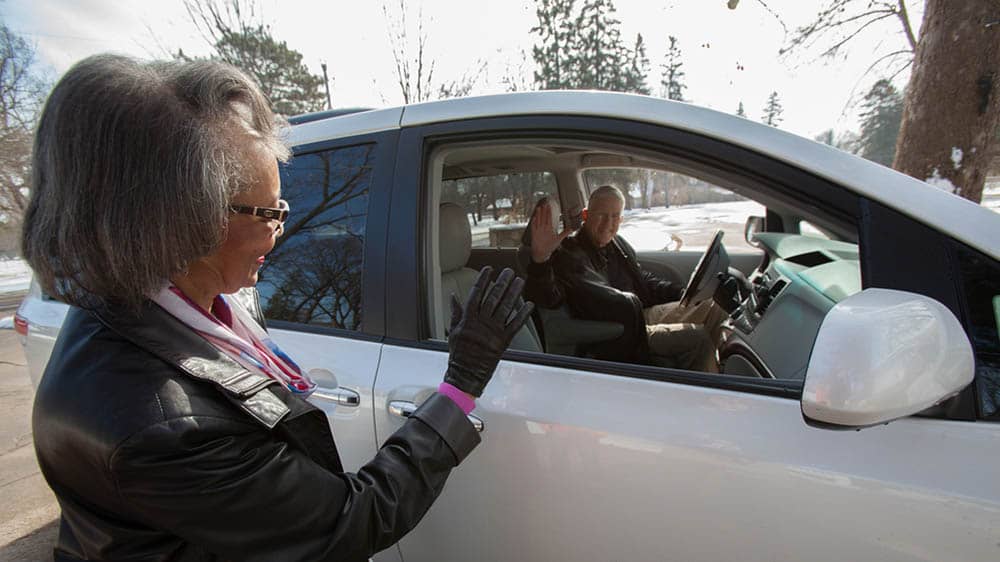 Senior female client waving at male Right at Home caregiver who just dropped her off outside in his white car, providing transportation services.