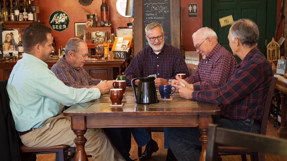 Four senior men and a male right at home caregiver sitting at a table in a coffee shop chatting