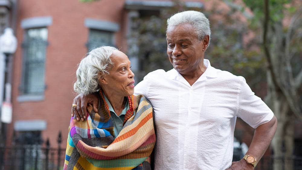 An elder senior couple with the man's arm around the woman, standing outside in front of brownstone building