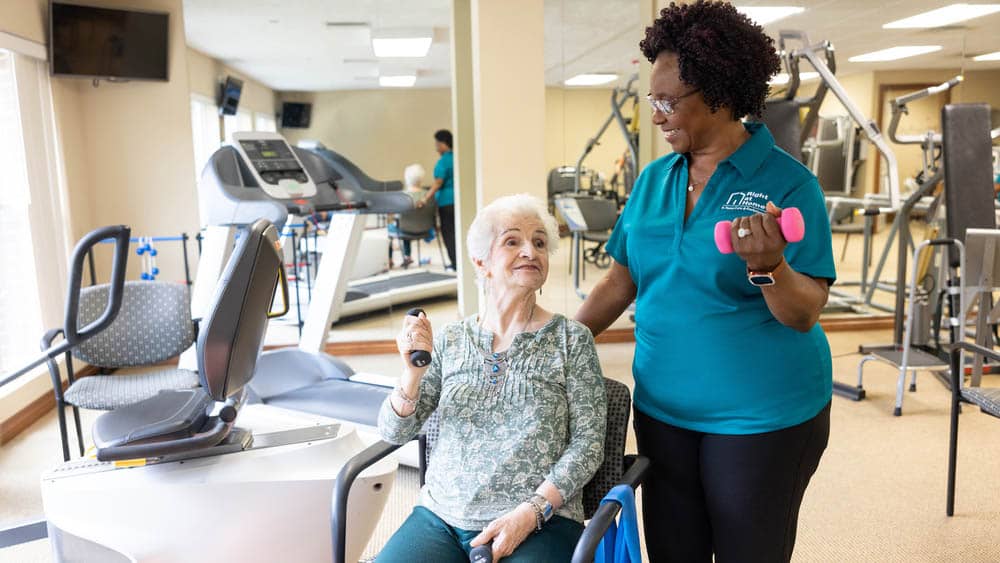 A senior female client and her female Right at Home caregiver are lifting small weights together inside of a small gym