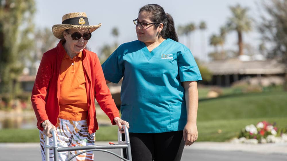 Senior female client using a walker while a female Right at Home caregiver walks beside her outside on a sunny day