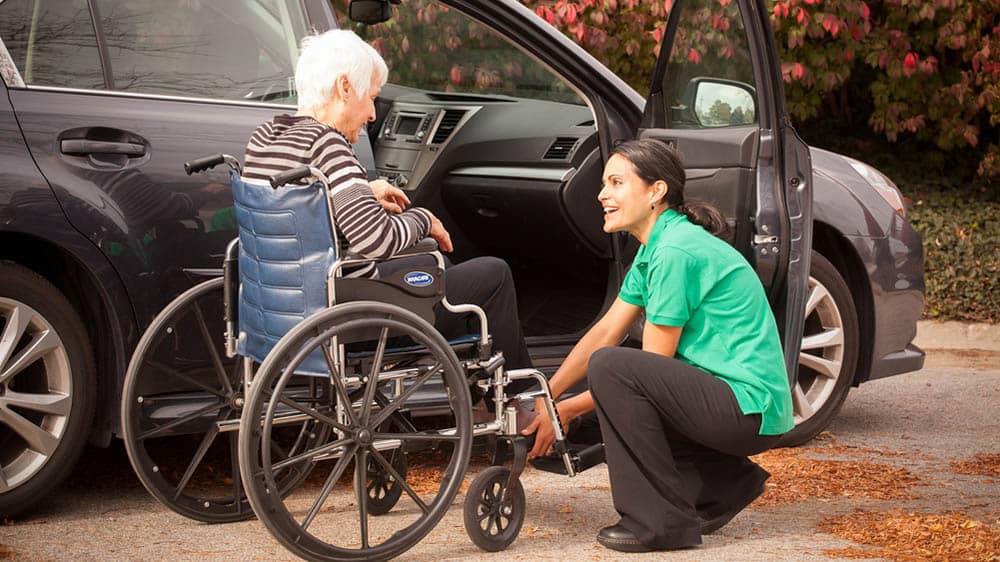 A female Right at Home caregiver is helping a senior female client into her wheelchair from the car outside