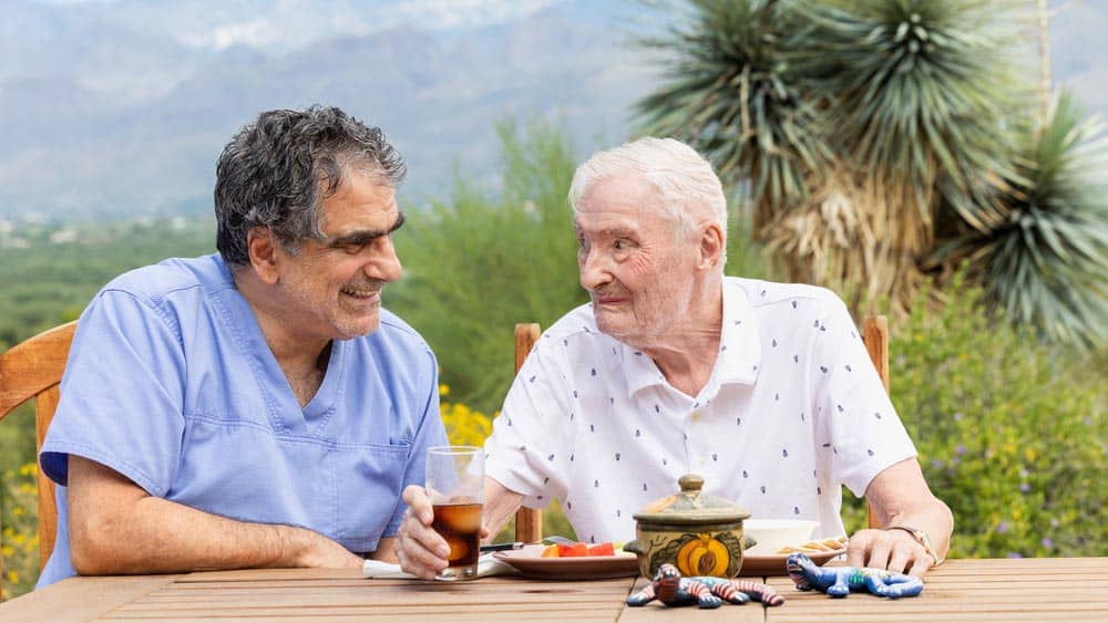Male Right at Home caregiver is sitting next to a senior male client who is enjoying a meal at an outside table