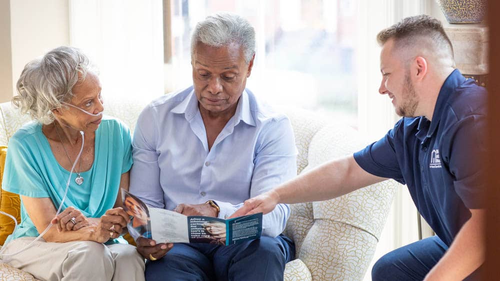 A male Right at Home staff member is sitting with a senior couple while they read a brochure during an in-home assessment visit