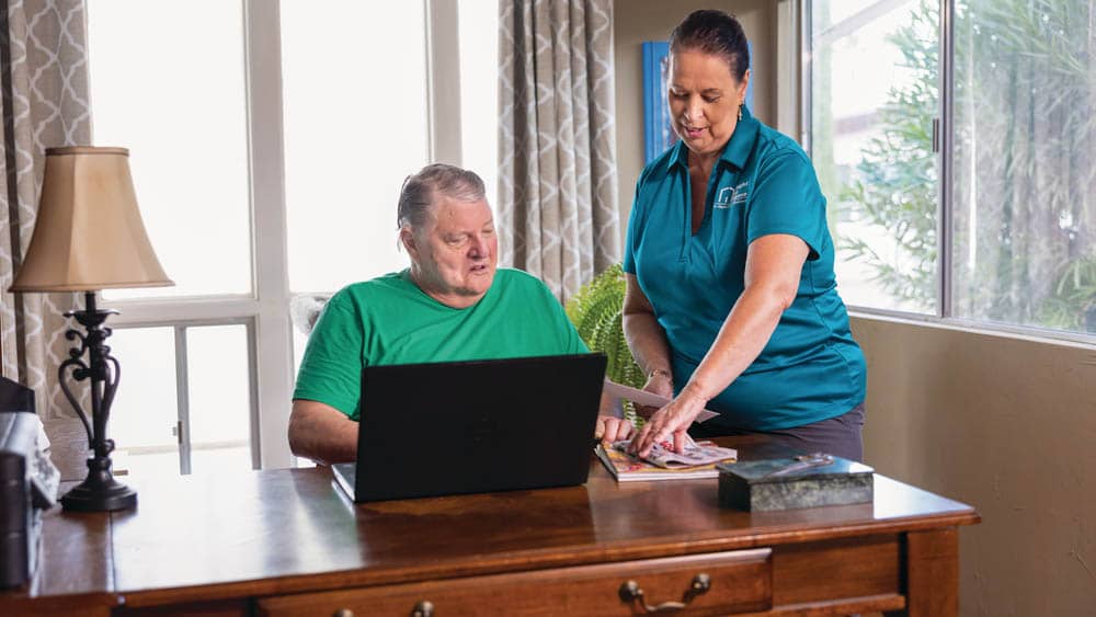 Senior male client sitting at a desk in front of a laptop computer while a female Right at Home caregiver stands near by