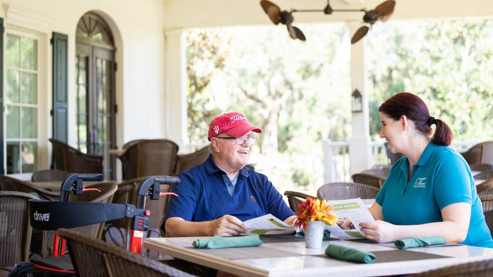 A senior male client is sitting at a table outside on a patio, his wheelchair on one side of him, his female Right at Home caregiver on the other.