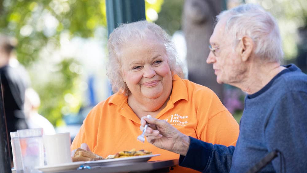 Female Right at Home caregiver look at a senior male client who is enjoying his lunch outside