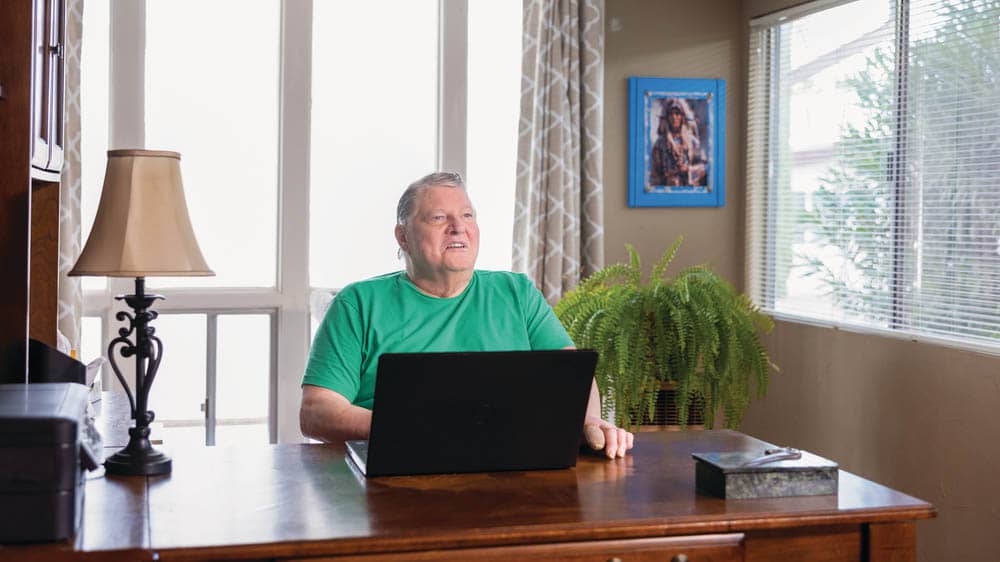 A senior male, sitting at a desk with a computer, in a home office