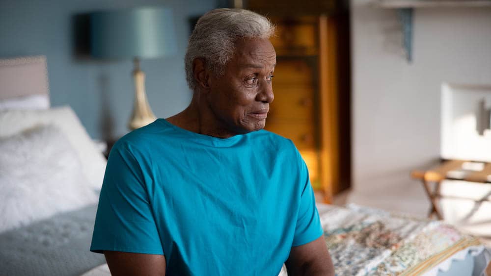 A senior male client sitting on the edge of a bed alone