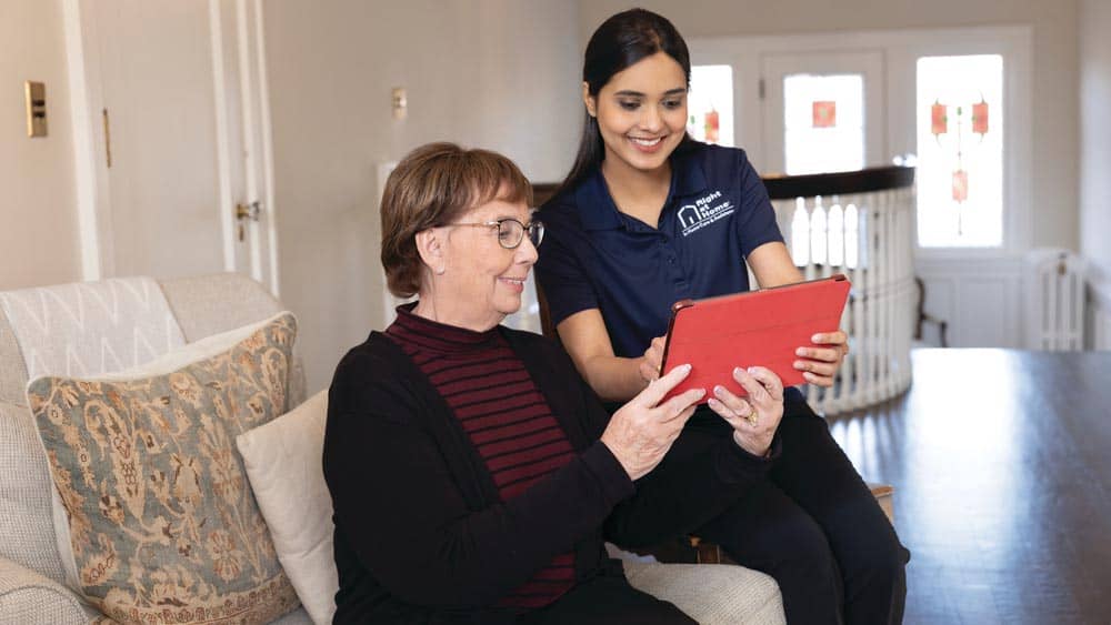 Female Right at Home caregiver sitting on the arm rest of a couch, helping a sitting female client sitting on the couch look at something on a tablet