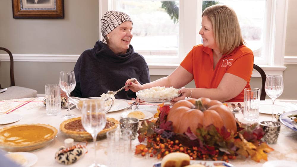 Female Right at Home caregiver and senior female client eating a Thanksgiving meal at the dining room table inside of client's home