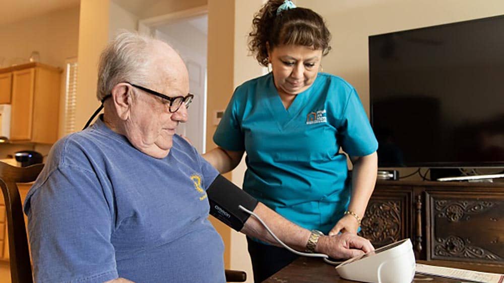 A female Right at Home caregiver is monitoring a senior male client take a blood pressure measurement 