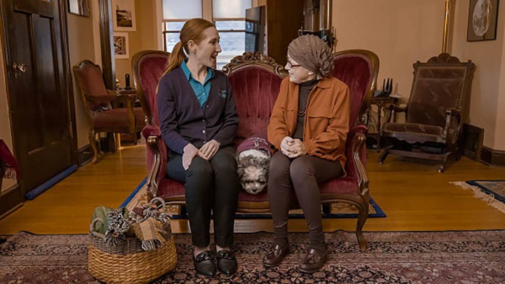 A senior female client is sitting on a couch with her female Right at Home caregiver, with a small dog laying down between them