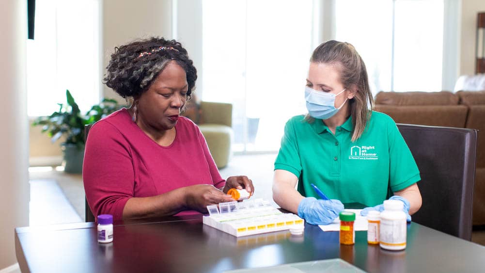 Female Right at Home caregiver supervising a senior female client sort out medication for the week into a planner