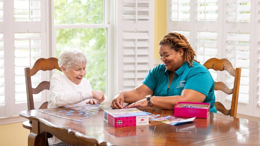 A senior female client is putting a puzzle together at the kitchen table with the assistance of a female Right at Home caregiver.