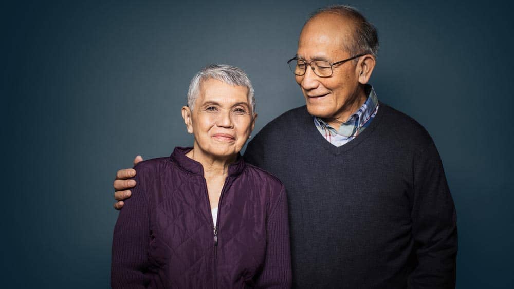 A senior man standing next to a senior female with his arm around her shoulder