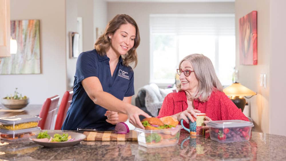 A female Right at Home caregiver helping prepare sandwiches at a kitchen counter while senior female client watches with a coffee.