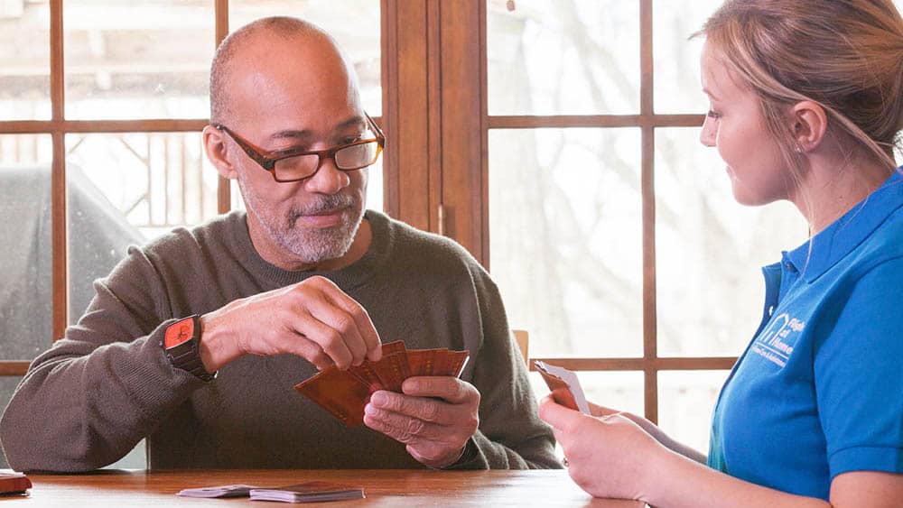 A senior aged man playing cards with a female Right at Home caregiver at a kitchen table.