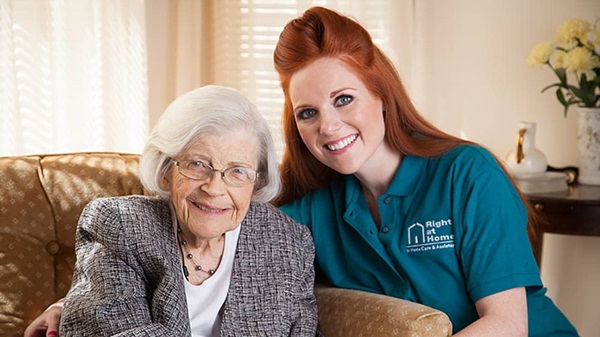 A female Right at Home caregiver smiling while posing next to a senior female sitting on a chair