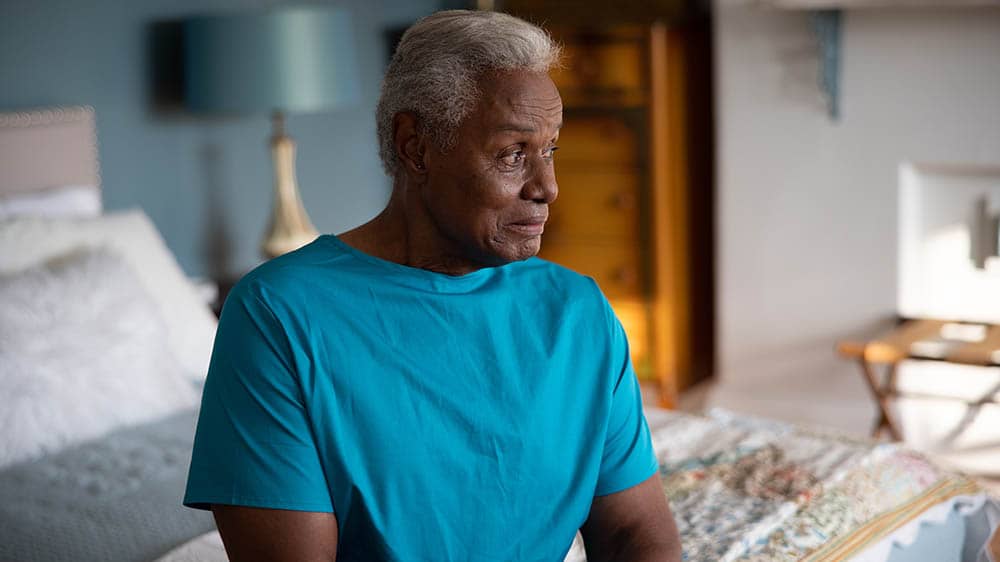 A senior male is sitting on the edge of his bed in his bedroom