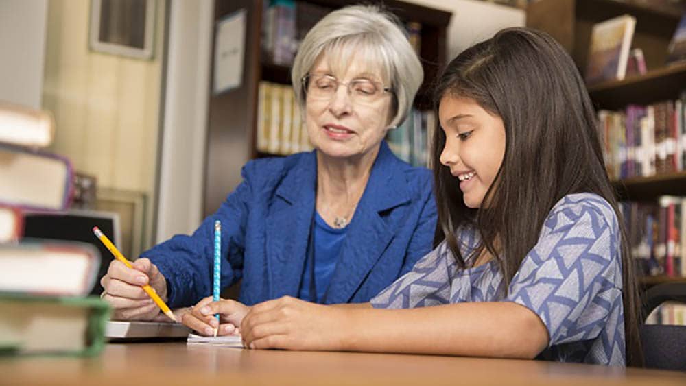 A senior female is teaching a young female child at a table in a library