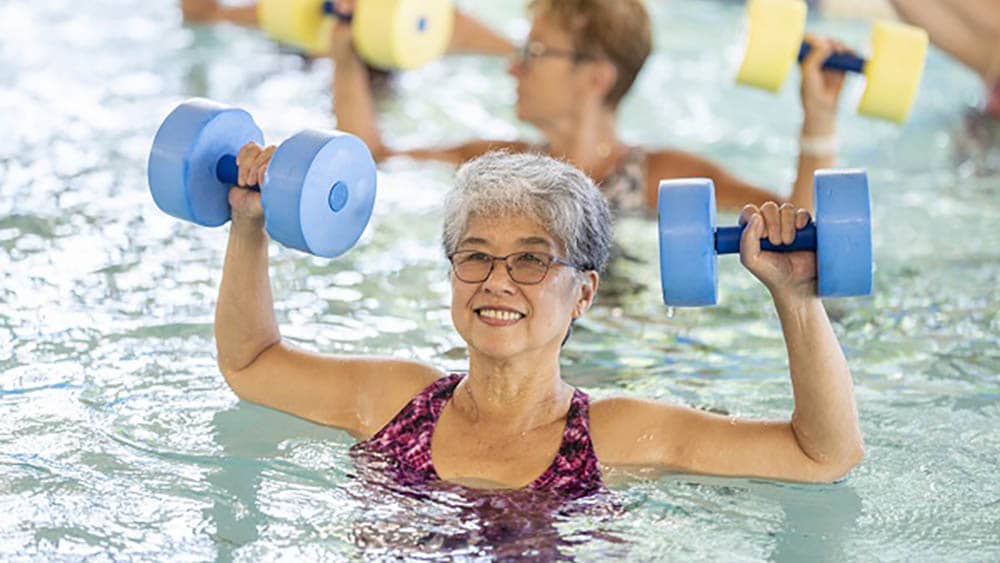 Senior female participating in water aerobics in a pool is holding foam weights 