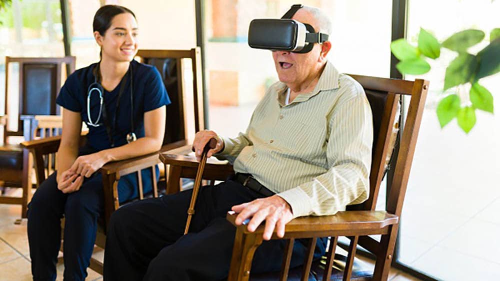 A senior male client sitting in a rocking chair is wearing virtual reality headset while his female Right at Home caregiver is sitting in the chair next to him observing. 