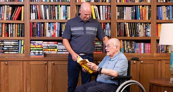 Senior man in a wheelchair reading a book in a library with his male caregiver