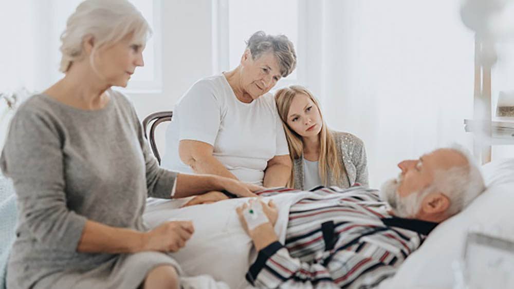 A senior man laying on a bed, surrounded by his concerned family.